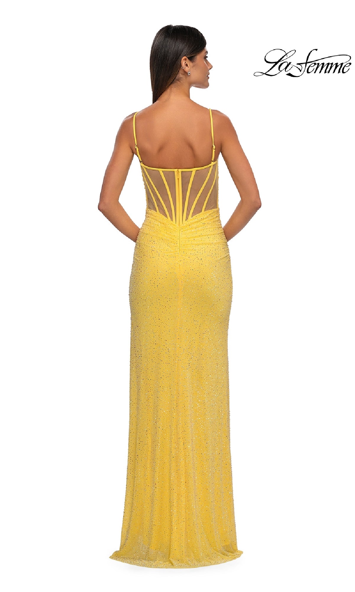 A woman in the La Femme 32578 long beaded prom dress, featuring a yellow hue, floor-length silhouette, sleeveless design with thin straps, and sheer-back detail, stands facing away from the camera.